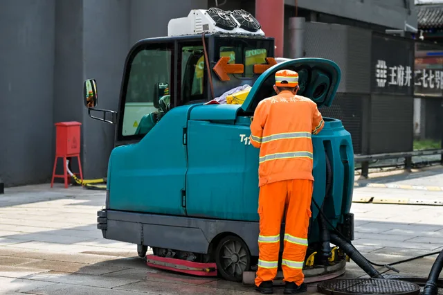 A street cleaner in uniform operates a cleaning machine on an urban sidewalk. Commercial cleaning equipment costs: new against used